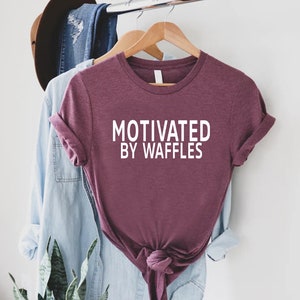 K&ouml;nnte beinhalten: Ein maronfarbenes T-Shirt mit dem Text "Motivated by Waffles" in wei&szlig;en Buchstaben.