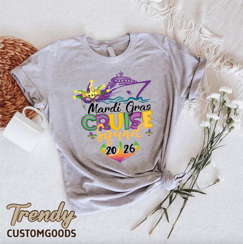 May include: A light grey short-sleeved t-shirt featuring a graphic of a cruise ship, a jester mask, and the text Mardi Gras CRUISE squad 20 26 in purple, green, and yellow. The design includes small fleur-de-lis accents and a colourful anchor icon.