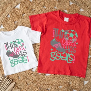 May include: Two white t-shirts with a green and pink graphic that says "This girl has goals" and a soccer ball and shoe design. One shirt is red and the other is white.