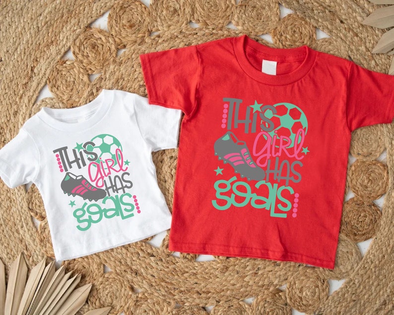May include: Two white t-shirts with a green and pink graphic that says "This girl has goals" and a soccer ball and shoe design. One shirt is red and the other is white.