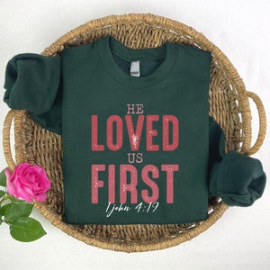 Puede incluir: Una sudadera verde oscuro con las palabras "He Loved Us First" en letras rojas y la referencia b&iacute;blica "1 Juan 4:19" en letras blancas m&aacute;s peque&ntilde;as.