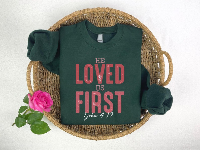 Puede incluir: Una sudadera verde oscuro con las palabras "He Loved Us First" en letras rojas y la referencia b&iacute;blica "1 Juan 4:19" en letras blancas m&aacute;s peque&ntilde;as.