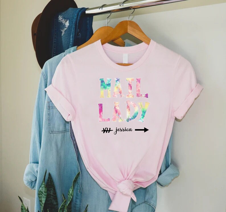 May include: Pink t-shirt with a tie-dye design that says "Mail Lady" with an arrow pointing to the right and the text "# Jessica" underneath.