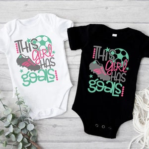 May include: Two baby onesies, one white and one black, with a soccer ball, cleats, and the text "This girl has goals!" printed on them.