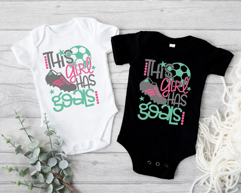 May include: Two baby onesies, one white and one black, with a soccer ball, cleats, and the text "This girl has goals!" printed on them.