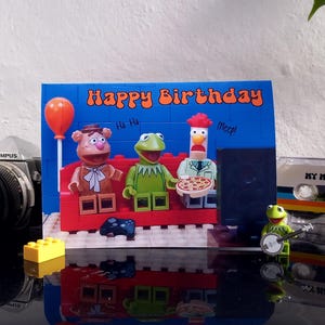May include: A birthday card with the words "Happy Birthday" and Lego figures of Kermit the Frog, Fozzie Bear, and a chicken. A vintage Olympus camera and a cassette tape are also visible.