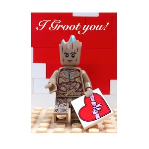 May include: A Lego figure of Groot from Guardians of the Galaxy holding a heart-shaped chocolate box with a pink ribbon. The background is red with the text "I Groot you!" in white.