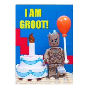 May include: A Lego Groot figure with an orange balloon stands beside a white birthday cake with blue ribbons. The background is a blue wall with the words "I AM GROOT!" in yellow.