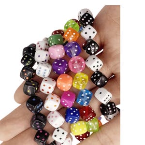 May include: A collection of small, cube-shaped beads designed to resemble dice, featuring various colours including black, white, red, orange, yellow, green, blue, and purple, each marked with contrasting dots representing standard numerical values.