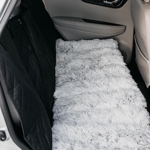 May include: A white fluffy car seat cover in the backseat of a car. The seat cover is covering the back seats and the floor of the car. The car seats are black.