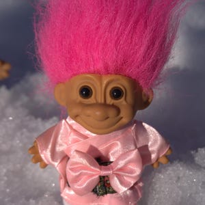 May include: A vintage Troll doll with a tan face, large black eyes, and a bright pink, spiky hair. The doll wears a pink satin outfit with a large bow and white fuzzy boots. A nostalgic collectible.