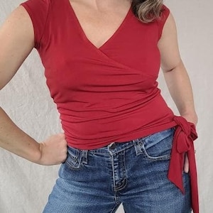 May include: A red wrap top with a V-neck and short sleeves. The top is tied at the waist with a long sash. The model is wearing blue jeans.