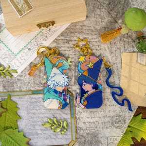 May include: Two rectangular acrylic keychains featuring anime-style characters in wizard hats. The left keychain shows a white-haired character in a light blue hat, while the right keychain depicts a dark-haired character in a dark blue hat with stars.