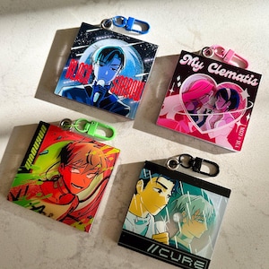 Pode incluir: Quatro chaveiros quadrados diferentes com ilustrações e texto em estilo anime coloridos. Os chaveiros apresentam um chaveiro preto e azul com um personagem cantando, um chaveiro rosa e branco com dois personagens se beijando, um chaveiro verde e vermelho com um personagem sorrindo e um chaveiro preto e verde com dois personagens se olhando.