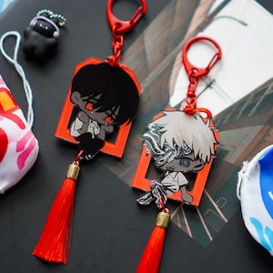 May include: Two anime-style keychains with red accents and tassels. Each keychain features a character in a red frame. One character has black hair, the other has white hair. Additional items include a small black figure and two decorative fish-shaped bags.