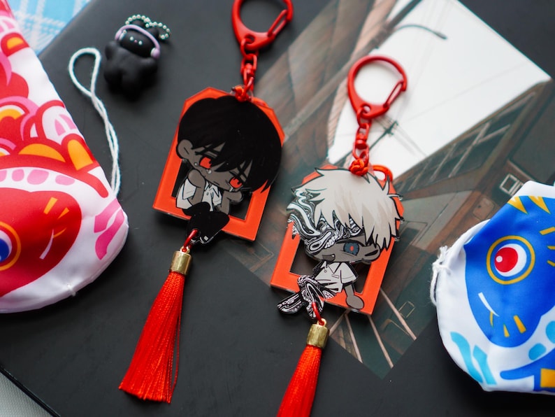 May include: Two anime-style keychains with red accents and tassels. Each keychain features a character in a red frame. One character has black hair, the other has white hair. Additional items include a small black figure and two decorative fish-shaped bags.