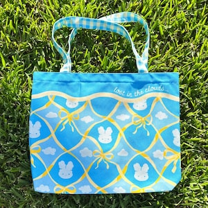 Puede incluir: Una bolsa de tela azul con un asa de cuadros blancos y un patrón de conejito blanco. La bolsa tiene el texto "lost in the clouds..." impreso en la parte delantera.