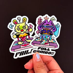 May include: Two die-cut stickers featuring cartoon rabbits on dance arcade platforms. The left sticker is yellow with the text FAIL, and the right sticker is purple with the text FULL COMBO. Both designs include colorful directional arrows.