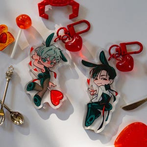 Könnte beinhalten: Zwei Acryl-Schlüsselanhänger mit Anime-Figuren mit Hasenohren, jeweils befestigt an einem roten Karabinerverschluss und einem kleinen roten Herzanhänger. Die Figuren tragen dunkelgrüne und schwarze Outfits mit kleinen roten Herzmotiven.