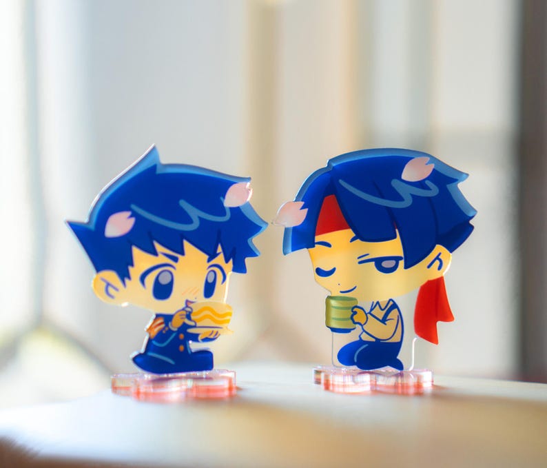 May include: Two acrylic anime-style figures. One figure is holding a bowl of ramen, the other is holding a cup. Both figures have blue hair, yellow skin, and are on clear bases with pink accents.