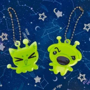 Könnte beinhalten: Zwei grüne Schlüsselanhänger mit Alien-Motiv und silbernen Kettenbefestigungen. Ein Schlüsselanhänger zeigt einen katzenartigen Alien mit einem lächelnden Gesicht, während der andere einen überraschten Ausdruck hat. Der Hintergrund ist dunkelblau mit einem Sternbildmuster und sternförmigen Akzenten.