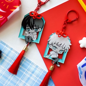 May include: Two anime-style character keychains with red tassels and a red clasp. The characters are in a teal frame. Other charms include a black bear with headphones, a white cloud, and a yellow star. The background is red and blue.
