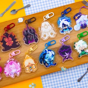 May include: A collection of colorful acrylic keychains featuring various cartoon characters. Each keychain has a unique design with vibrant colors and attached to a metal clasp. The keychains are arranged on a wooden cutting board.