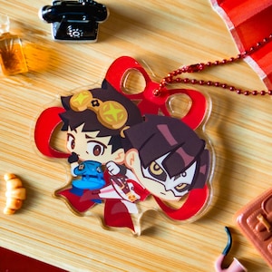 May include: A clear acrylic charm featuring two anime-style characters with brown and purple hair, wearing costumes. The charm has a red ribbon and a red beaded chain. Other miniature items include a magnifying glass, a croissant, and a miniature telephone.