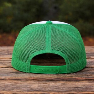 K&ouml;nnte beinhalten: Eine gr&uuml;ne und wei&szlig;e Trucker-Kappe mit Mesh-R&uuml;ckseite und gebogenem Schirm. Die Kappe hat einen verstellbaren gr&uuml;nen Druckknopfverschluss und eine wei&szlig;e Frontplatte. Auf einer Holzoberfl&auml;che platziert.