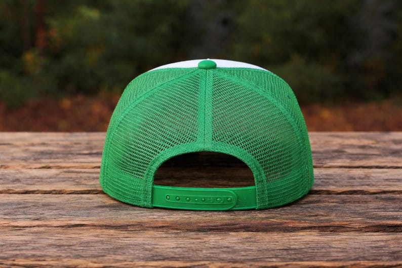 K&ouml;nnte beinhalten: Eine gr&uuml;ne und wei&szlig;e Trucker-Kappe mit Mesh-R&uuml;ckseite und gebogenem Schirm. Die Kappe hat einen verstellbaren gr&uuml;nen Druckknopfverschluss und eine wei&szlig;e Frontplatte. Auf einer Holzoberfl&auml;che platziert.