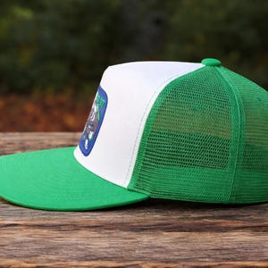 K&ouml;nnte beinhalten: Eine gr&uuml;ne und wei&szlig;e Trucker-M&uuml;tze mit gr&uuml;nem Schirm und Mesh-R&uuml;ckseite. Die M&uuml;tze hat einen Aufn&auml;her mit einem Design auf der Vorderseite. Die M&uuml;tze liegt auf einer Holzoberfl&auml;che.