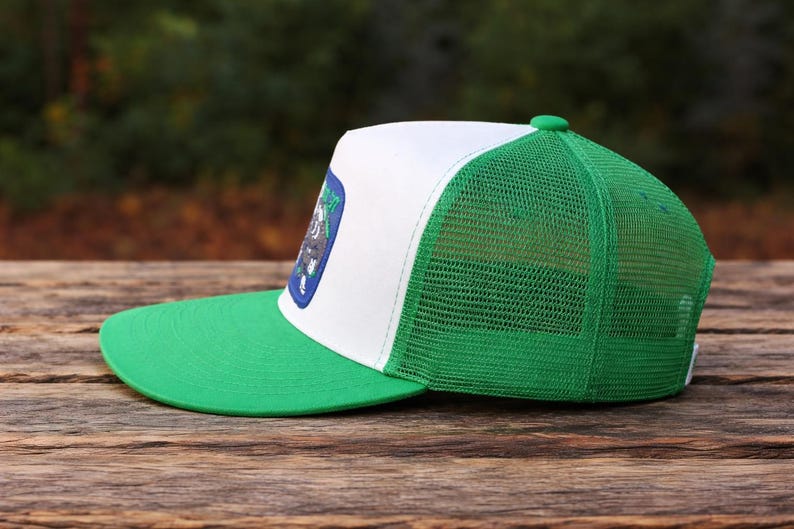 K&ouml;nnte beinhalten: Eine gr&uuml;ne und wei&szlig;e Trucker-M&uuml;tze mit gr&uuml;nem Schirm und Mesh-R&uuml;ckseite. Die M&uuml;tze hat einen Aufn&auml;her mit einem Design auf der Vorderseite. Die M&uuml;tze liegt auf einer Holzoberfl&auml;che.