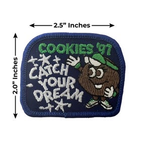 K&ouml;nnte beinhalten: Ein marineblauer, gestickter Aufn&auml;her, 6,35 cm breit und 5,08 cm hoch, mit dem Text "COOKIES '97" und "CATCH YOUR DREAM" in Wei&szlig; und Gr&uuml;n. Er zeigt eine Cartoon-Keksfigur mit gr&uuml;nem Hut und Turnschuhen.
