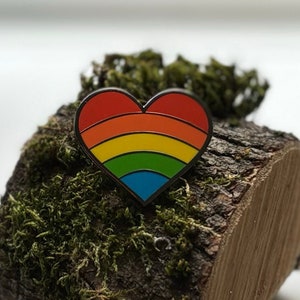 May include: A rainbow heart-shaped enamel pin on a piece of wood covered in moss.