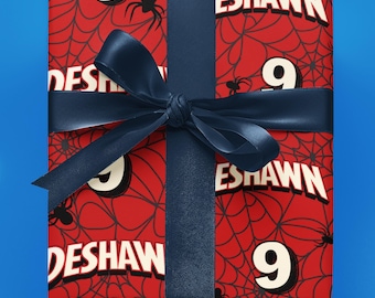 May include: A rectangular gift box wrapped in red paper featuring a black spiderweb pattern, small black spiders, the number 9, and the name DESHAWN in white lettering. The package is tied with a dark blue satin ribbon centered horizontally.