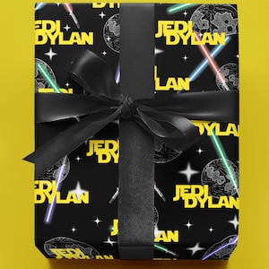 Op de afbeelding: Een ingepakt cadeau met zwart cadeaupapier met een herhaald patroon van planeten, lichtzwaarden en de tekst "Jedi Dylan" in geel. Een zwart lint en strik beveiligen het cadeau, tegen een gele achtergrond.