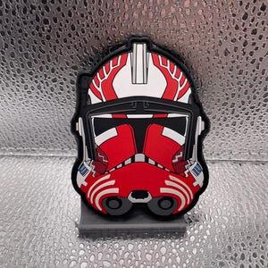May include: A red and white Star Wars clone trooper helmet patch. The helmet has a red and white paint scheme with a black visor. The patch is made of a durable material and is perfect for adding a touch of Star Wars to your gear.