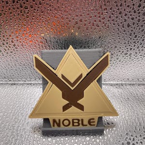 May include: A 3D printed stand with a beige triangle shape and a brown symbol. The stand has the word "NOBLE" printed on the base.