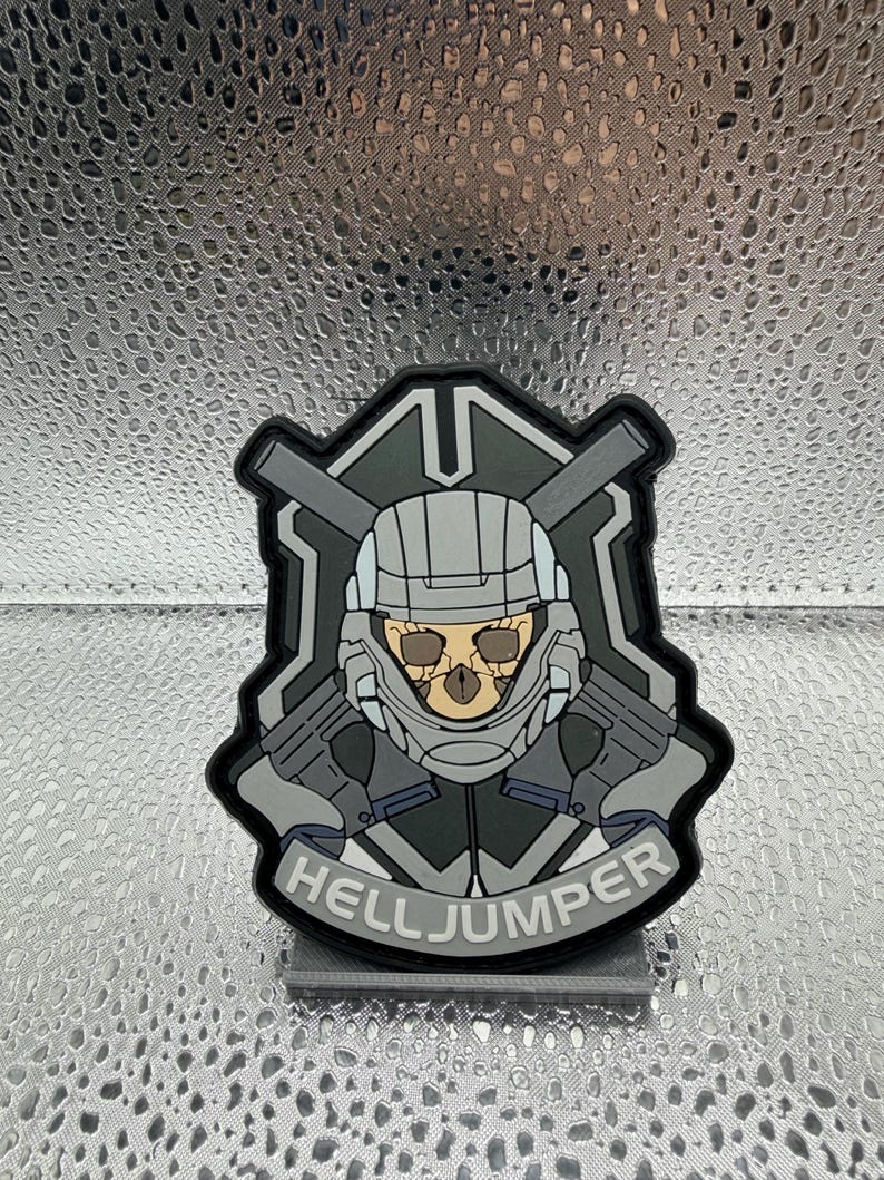 May include: A black, gray, and white patch featuring a helmeted skull with crossed rifles and the text "HELLJUMPER". The patch has a military or sci-fi theme and is likely a collectible or accessory.