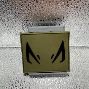 May include: An olive green patch with a black stylized eye design. The patch is rectangular and appears to be made of fabric. The background is a textured, silver material.