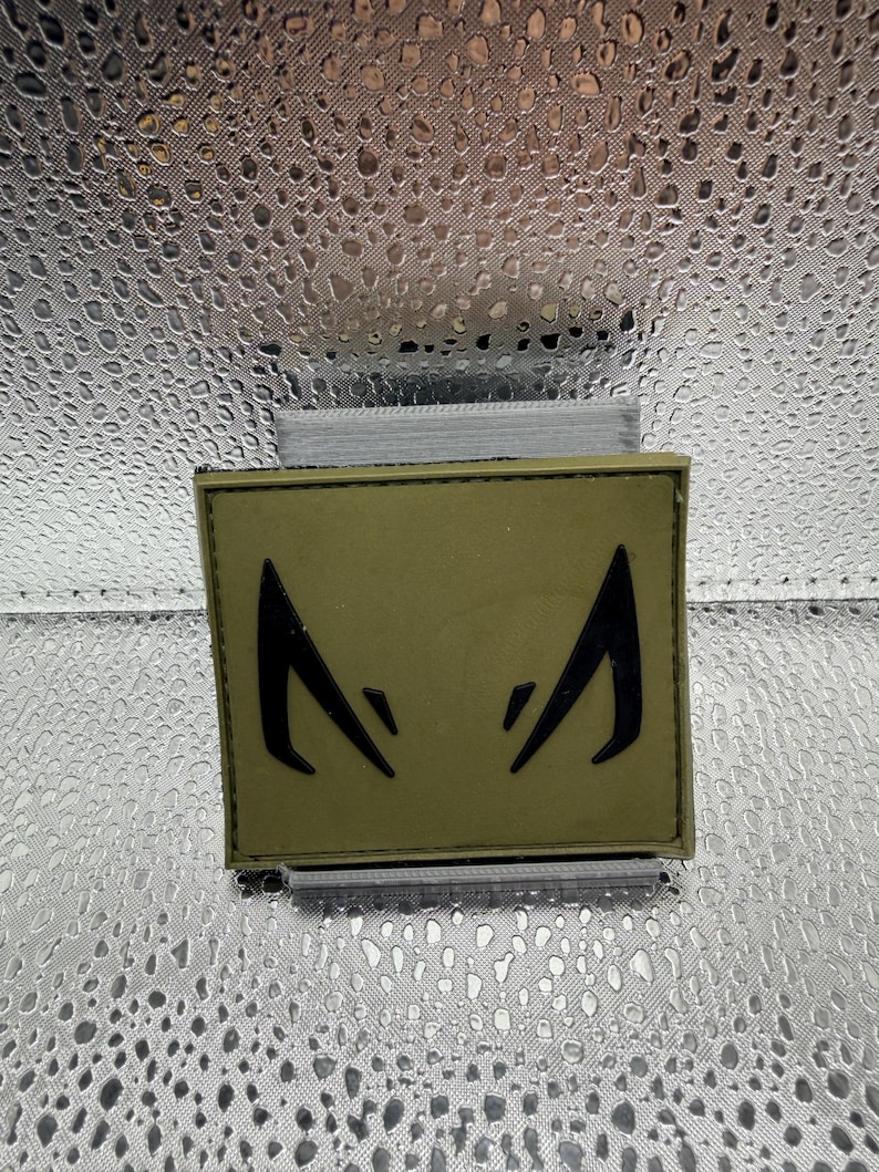 May include: An olive green patch with a black stylized eye design. The patch is rectangular and appears to be made of fabric. The background is a textured, silver material.