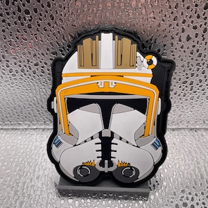May include: A patch featuring a white and gray helmet with orange and yellow accents, resembling a clone trooper helmet. The patch has a black border and is set against a textured silver background.
