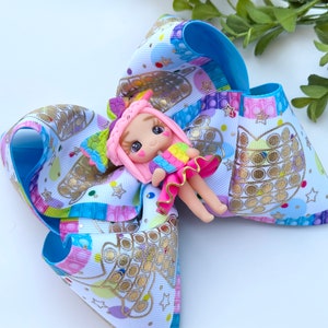 May include: A blue, pink, and white bow with a gold pop-it design and a small pink doll with rainbow hair and a rainbow dress.