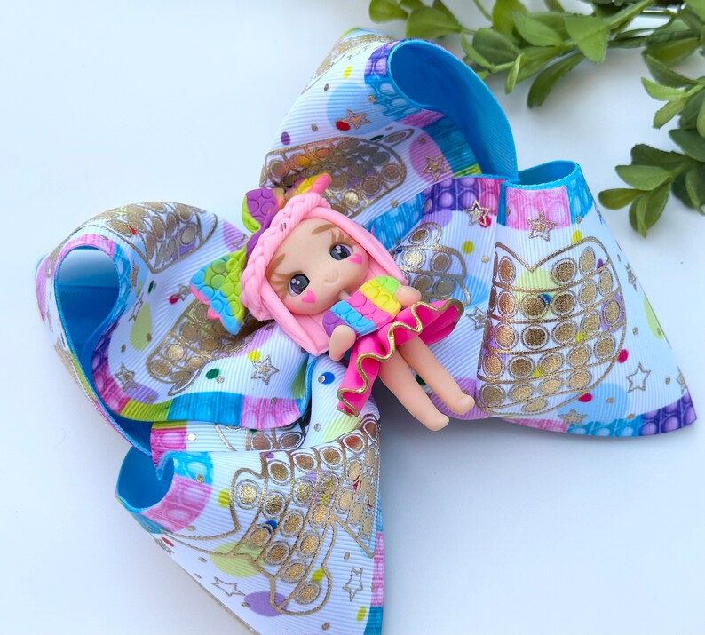 May include: A blue, pink, and white bow with a gold pop-it design and a small pink doll with rainbow hair and a rainbow dress.