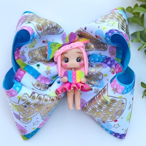 May include: A colorful hair bow with a white background and a pop-it pattern. A small pink doll with rainbow hair and a pop-it heart is attached to the center of the bow.