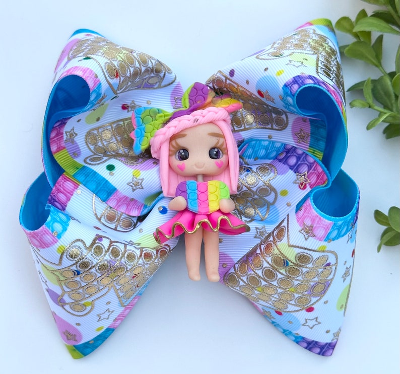 May include: A colorful hair bow with a white background and a pop-it pattern. A small pink doll with rainbow hair and a pop-it heart is attached to the center of the bow.