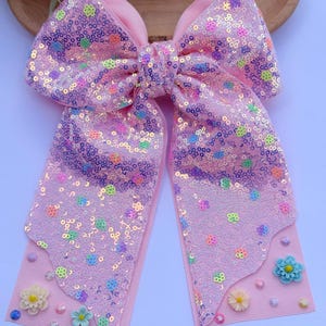 May include: A large pink hair bow with a sequined design and floral embellishments. The bow features a double-layered design with a solid pink ribbon base and a sequined overlay. Small, colorful flower accents are attached to the bottom of the bow.