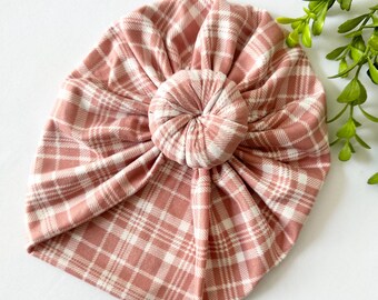 May include: A pink and white plaid baby turban hat. The hat is made of soft fabric and features a knotted design at the top. The plaid pattern is a classic design, and the hat is likely for infants or toddlers.