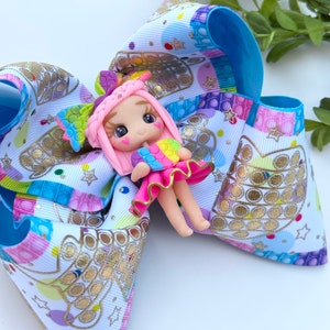 May include: A colorful hair bow with a pop it design and a pink doll with rainbow hair and a rainbow pop it in her hand.