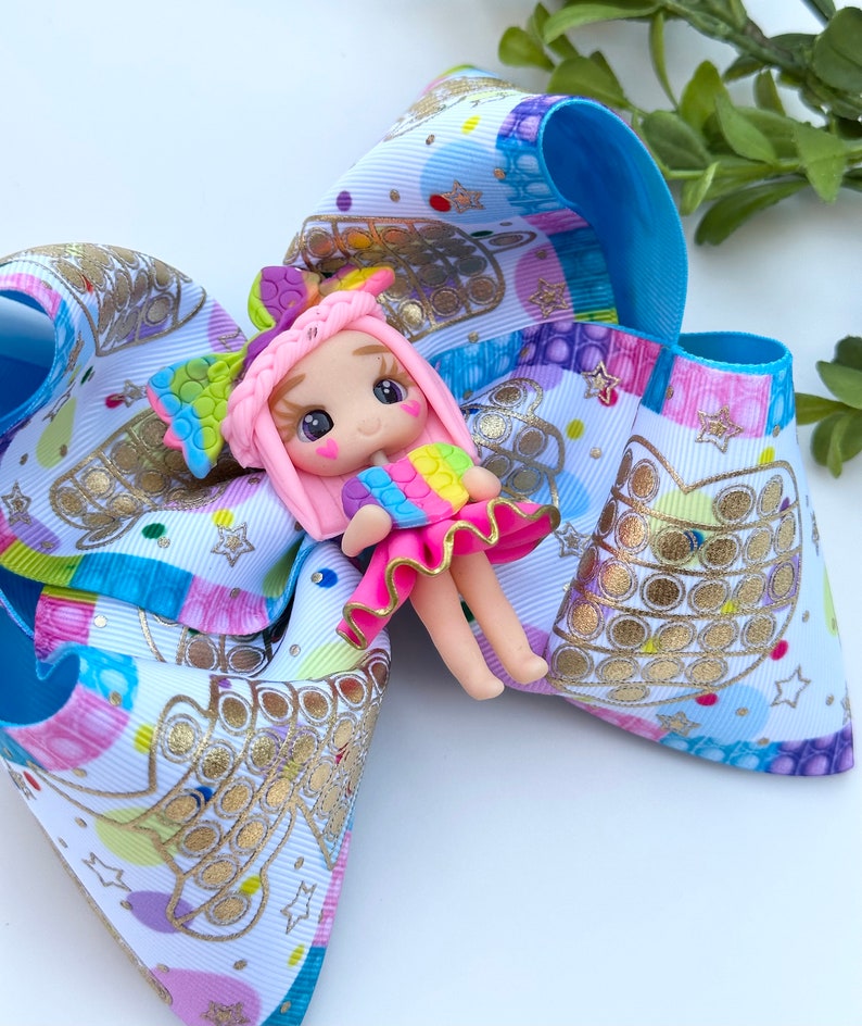 May include: A colorful hair bow with a pop it design and a pink doll with rainbow hair and a rainbow pop it in her hand.